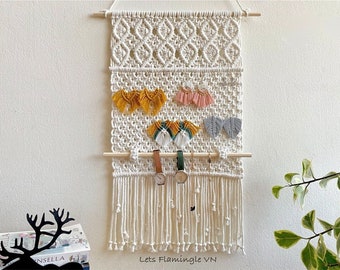 May include: An off-white macrame wall hanging organiser featuring a diamond-patterned top section, a grid-like middle for earrings, and a wooden dowel for watches and bracelets. The piece ends in long fringe and displays the text Lets Flamingle VN.