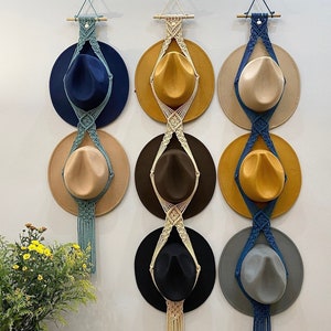 Könnte beinhalten: Drei Makramee-Huthalter mit jeweils drei Hüten. Die Halter sind aus blauem und weißem Kordel gefertigt und die Hüte sind braun, beige und blau.