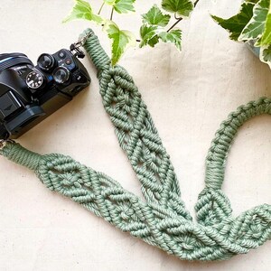 May include: A sage green macrame camera strap featuring an intricate knotted pattern with a repeating diamond design. The strap is attached to a black digital camera with silver metal swivel clips at both ends, showcasing a textured cord construction.