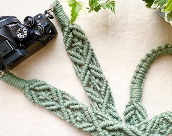 May include: A sage green macrame camera strap featuring an intricate knotted pattern with a repeating diamond design. The strap is attached to a black digital camera with silver metal swivel clips at both ends, showcasing a textured cord construction.