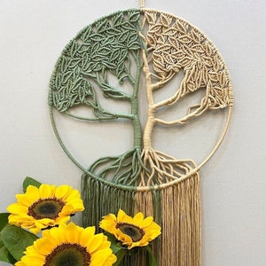 May include: A macrame wall hanging in the shape of a tree with green and beige yarn. The tree has a thick trunk and branches with leaves. The roots are visible at the bottom of the tree. The wall hanging is hanging on a white wall. There are sunflowers in the foreground.