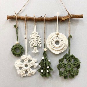 Può includere: Una collezione di ornamenti natalizi in macramè fatti a mano in bianco e verde, sospesi da un ramo di legno naturale. Gli ornamenti includono una ghirlanda, un fiocco di neve, un albero di Natale e disegni circolari, tutti con dettagli di annodatura.