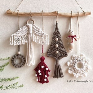 May include: A set of five handmade macrame ornaments hanging from a wooden branch. The ornaments include a white snowflake, a brown Christmas tree, a white angel wing, a red and white tassel, and a brown ring. The ornaments are all made with natural fibers and have a rustic, bohemian style. The text "Lets Flamingle VN" is visible at the bottom of the image.