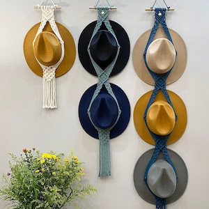 May include: Three macrame hat holders with different colored hats. The holders are made of white and blue cord and have a wooden dowel at the top. The hats are brown, beige, and gray.