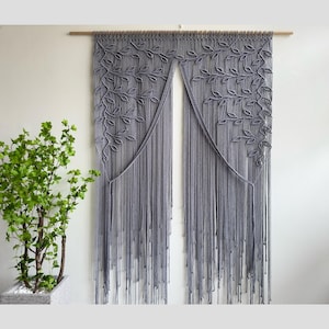 Pode incluir: Uma tapeçaria de parede cinza em macramê com um desenho de folhas. A peça de arte têxtil é suspensa por uma vara de madeira e apresenta fios longos e com franjas. À esquerda, há uma planta em vaso com folhas verdes.