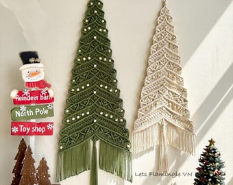 May include: Two wall-hanging macrame Christmas trees, one in olive green and one in cream, featuring intricate knotted patterns and wooden beads. Beside them is a signpost with text reading Reindeer barn, North Pole, and Toy shop.