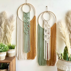 May include: Two macrame wall hangings with wooden hoops and long, fringed strands. One features cream, sage green, and mustard yellow, the other cream, sage green, and mustard yellow. Both have leaf patterns.