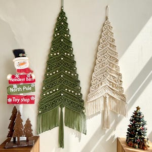 May include: Two macrame Christmas tree wall hangings, one green and one cream, with fringe at the bottom. The green tree has small white lights. A snowman sign that reads "Reindeer barn", "North Pole", and "Toy shop" is also visible.