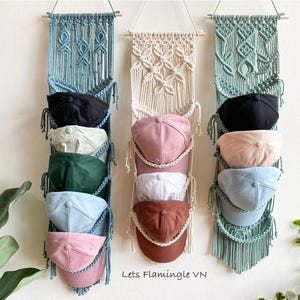 Puede incluir: Tres organizadores de sombreros de macramé en azul, crema y verde, cada uno con varias gorras de béisbol. Las gorras son de varios colores, incluyendo negro, rosa, blanco y azul. Los organizadores están colgados en una pared blanca.