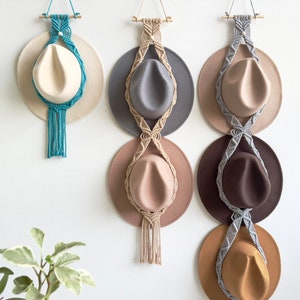 May include: Three macrame hat holders with different coloured tassels hanging on a white wall. Each holder has a round base and is designed to hold a hat. The hats are different colours and styles.