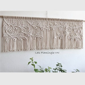 Pode incluir: Uma tapeçaria de macramé bege com um design de folhas. A tapeçaria é feita com cordão de algodão natural e tem franjas na parte inferior. O texto "Lets Flamingle VN" é visível na parte inferior da imagem.