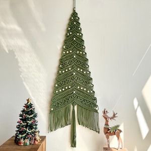 May include: A green macrame Christmas tree wall hanging with white bead accents. The tree has a fringe at the bottom and is hanging on a white wall. A small decorated Christmas tree and a wooden reindeer are also in the image.