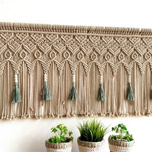 May include: A macrame wall hanging with a geometric pattern and tassels. The hanging is made of natural beige cord and has white beads and green tassels.