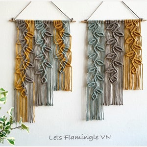 Op de afbeelding: Twee macramé wandkleden met bladerpatronen in tinten grijs, mosterdgeel en blauwgroen. De wandkunst is opgehangen aan een houten deuvel. De tekst "Lets Flamingle VN" is zichtbaar onderaan de afbeelding.