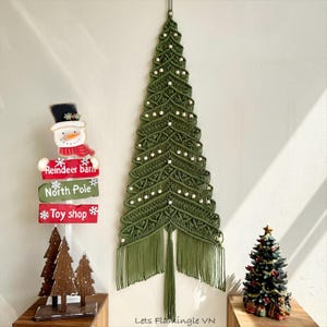 May include: A wall-hanging macrame Christmas tree in olive green featuring intricate knotted patterns and small wooden beads. Beside it stands a snowman signpost labeled Reindeer barn, North Pole, and Toy shop, plus a small decorative tree.