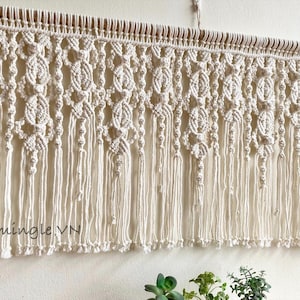May include: A cream-coloured macrame wall hanging suspended from a wooden dowel, featuring intricate knotted patterns, repeating oval shapes, and long vertical fringe. The text "Lets Flamingle VN" is printed in the lower left corner of the image.