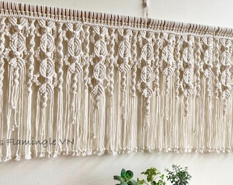 May include: A cream-colored macrame wall hanging suspended from a wooden dowel, featuring intricate knotted patterns, repeating oval shapes, and long vertical fringe. The text "Lets Flamingle VN" is printed in the lower left corner of the image.