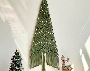 May include: A wall hanging in the shape of a Christmas tree made of olive green macrame cord with white wooden beads integrated into the knotting pattern. The piece features a long fringe at the base and the text Lets Flaningle VN at the bottom.
