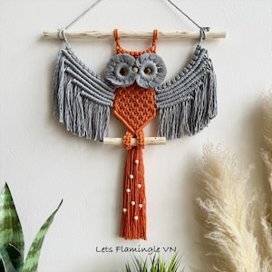 May include: A macrame owl wall hanging featuring an orange knotted body, grey fringed wings, and two large grey circular eyes with brown beads. The owl is suspended from a light-colored wooden dowel with a string and includes the text Lets Flamingle VN.