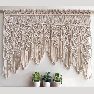 May include: A beige macrame wall hanging with a leaf design. The handmade textile features intricate knotting and a scalloped bottom edge. Three small potted plants sit below the macrame.