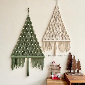 May include: Two triangular macrame wall hangings shaped like Christmas trees, one in olive green with white beads and one in off-white. Below them, a wooden signpost reads North Pole, Santa's House, and Workshop, with small figurines.