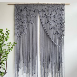 May include: A grey macrame wall hanging suspended from a wooden rod, featuring an intricate pattern of knotted leaves and vines that transition into long, vertical fringe strands that hang down to create a textured, layered appearance.