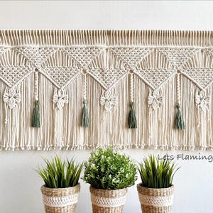 May include: A cream-coloured macrame wall hanging featuring a series of repeating triangular patterns, intricate knotted designs, and sage green tassels suspended from a wooden rod. The text "Lets Flamingle VN" is visible in the bottom right corner.