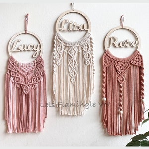 May include: Three macrame dreamcatchers with wooden hoops and personalized names, "Lucy", "Lisa", and "Rose". The dreamcatchers are made with natural cotton cord and feature a variety of knots and patterns. The dreamcatchers are hanging from a white wall.