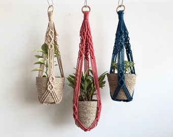 May include: Three macrame plant hangers with different colored cords, each holding a small potted plant. The hangers are made of natural fiber rope and have a simple, minimalist design. The plants are green and leafy.