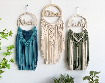 May include: Three macrame wall hangings with wooden name cutouts. The first is blue with the name "Kevin", the second is cream with the name "Lisa", and the third is green and cream with the name "Emily". Each piece is suspended by a string.