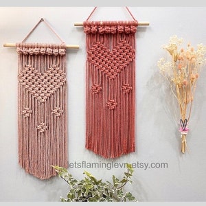 May include: Two pink macrame wall hangings with heart designs. The hangings are made with cotton cord and have a wooden dowel at the top. The hangings are displayed on a white wall with dried flowers and greenery.