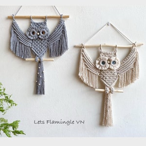 May include: Two macrame owl wall hangings, one in grey and one in beige, with wooden dowels and beaded tails. The owls have large eyes and are hanging on a white wall. Lets Flamingle VN is written below the owls.
