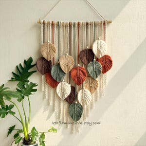 May include: A macrame wall hanging featuring a wooden dowel with hanging cream-colored cords and multiple leaf-shaped fiber ornaments in shades of rust, sage green, dark brown, and cream, with the text letsflaminglevn.etsy.com at the bottom.