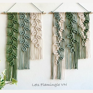 May include: Two macrame wall hangings featuring a leaf design in shades of green and cream. The hangings are crafted from cotton cord and are suspended from a wooden dowel.