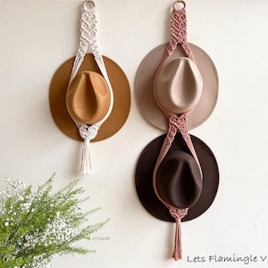 Puede incluir: Dos colgadores de macramé para sombreros hechos de cuerda, uno blanco y otro rosa empolvado, cada uno con un anillo de madera en la parte superior y una borla en la inferior. El blanco sostiene un sombrero canela, el rosa uno beige y otro marrón.
