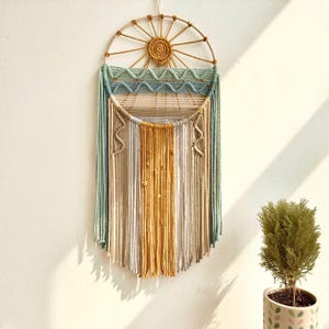 May include: A macrame wall hanging with a circular wooden frame and a sun design. The hanging features a variety of colors, including beige, light blue, and yellow.