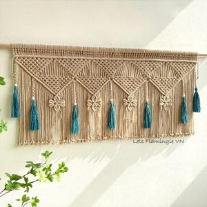 Op de afbeelding: Een beige macramé wandkleed met een reeks driehoekige patronen, ingewikkeld knoopwerk en decoratieve bloemvormige accenten. Het stuk is afgewerkt met meerdere blauwgroene kwastjes en kleine witte kralen, opgehangen aan een houten roede.