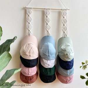 May include: A white macrame wall hanging with three rows of hooks for hanging baseball caps. The caps are in various colours, including pink, brown, black, green, blue, and white. The wall hanging is a great way to organise and display your favourite caps.