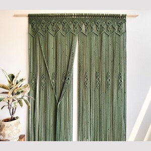 Op de afbeelding: Saliegroen macramé gordijn met ingewikkelde geometrische patronen aan de bovenkant en langs de verticale strengen. Het gordijn hangt aan een natuurlijke houten roede en voegt een vleugje bohemien stijl toe aan elk interieur.