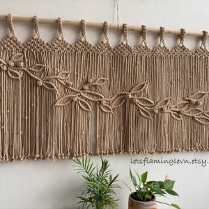 May include: A beige macrame wall hanging suspended from a wooden dowel, featuring a knotted leaf pattern across the centre and vertical fringe with small knots. The text letsflaminglevn.etsy.com is printed in the lower right corner.