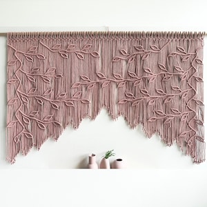 Op de afbeelding: Een roze macramé wandhanger met een bladontwerp. De hanger is gemaakt van katoenen koord en is bevestigd aan een houten lat.