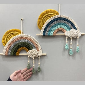 May include: Two macrame rainbow wall hangings with a sun and cloud design. The top rainbow is blue, green, and beige. The bottom rainbow is green, yellow, and beige. Both rainbows have a cloud with raindrops hanging from them.