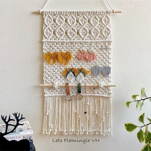 Peut inclure: Une suspension murale en macramé blanche avec une tringle en bois et des franges. La suspension murale a un motif géométrique et est décorée de boucles d'oreilles et de montres colorées. Le texte "Lets Flamingle VN" est imprimé en bas de la suspension murale.