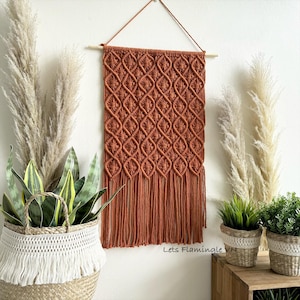 May include: A rectangular macrame wall hanging in a deep rust orange colour featuring a repeating diamond knot pattern and long fringe at the bottom. The piece is suspended from a light wooden dowel with a thin string for hanging on a wall.