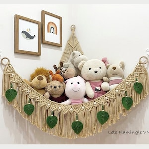 May include: A beige macrame toy hammock featuring a knotted cord design, wooden rings at the ends, and hanging green fabric leaf accents. The hammock is filled with various plush stuffed animals, including a lion, bear, and pig, against a wall.