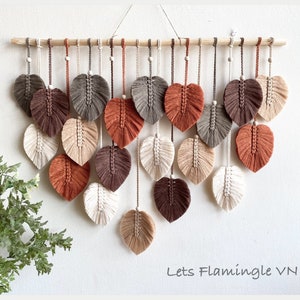 May include: A macrame wall hanging with 18 leaf-shaped decorations in shades of brown, beige, and white, suspended from a wooden dowel. Each leaf has a detailed, textured design. The wall decor is displayed against a white wall.