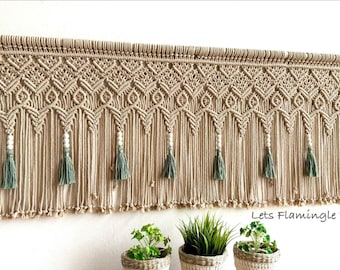 May include: A beige macrame wall hanging featuring intricate knotted patterns, white wooden beads, and sage green tassels suspended from a wooden dowel. The text "Lets Flamingle VN" is printed in the lower right corner of the image.