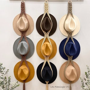 May include: A wall-mounted hat rack with six hats hanging from it. The rack is made of macrame and has a brown, beige, and cream colour scheme. The hats are in various colours, including brown, beige, cream, blue, and yellow.