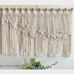 May include: A white macrame wall hanging with a leaf design. The hanging is made of cotton cord and has a wooden dowel at the top. The hanging is approximately 30 inches wide and 20 inches tall.