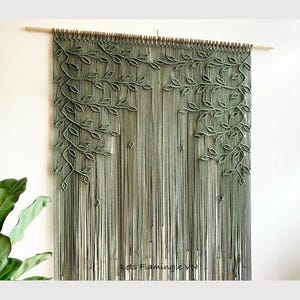 May include: A macrame wall hanging featuring a design of leaves and branches in shades of green. The artwork is suspended from a wooden dowel. The piece has a textured, layered appearance, creating a natural, organic aesthetic.